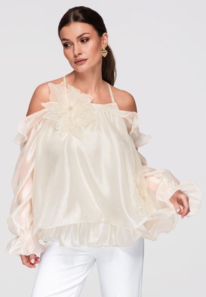 Mujer con cabello oscuro en coleta, vistiendo una blusa crema transparente con escote fuera del hombro y un gran detalle floral, y pantalones blancos sobre fondo gris.