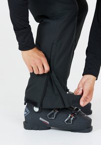 Zwarte skibroek met ritsen en klitteband aan de boorden, die wordt aangepast over een zwarte skischoen met een gestructureerd oppervlak en gespen, op een witte achtergrond.