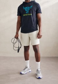 Grå kortärmad hoodie med blå och grön grafik, beige texturerade shorts, grå sportskor och vita strumpor som håller i ett hopprep.