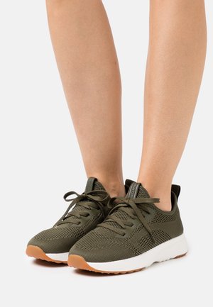 Pieds portant des baskets tricotées vert olive avec semelles blanches et bouts en caoutchouc couleur gomme, lacées et posées sur une surface blanche.