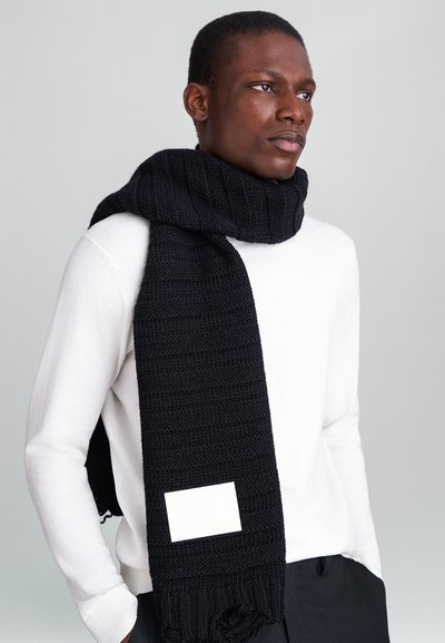 Musträne sall mustriga tekstuuriga, millel on ristkülikukujuline valge silt. Kandsime kaela ümber, valge sviitri peal.