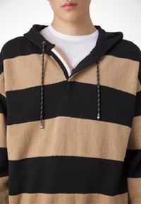 Jeune personne portant un sweat à capuche tricoté rayé large noir et marron clair avec un t-shirt blanc à col rond en dessous.