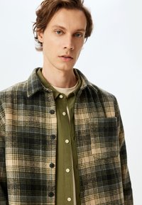 Chemise en flanelle à carreaux beige, vert olive et bleu marine, avec une fermeture à boutons sur le devant, un col et une poche poitrine simple. Texture douce et coupe décontractée.