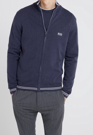 Mann trägt marineblauen Reißverschluss-Pullover mit Hugo Boss-Logo auf der Brust und graue Nadelstreifenhose, Hände in den Taschen, vor einfachem Hintergrund.