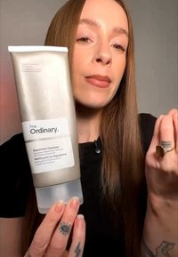 Femme aux longs cheveux tenant un tube de The Ordinary Squalane Cleanser, montrant le devant du produit et portant des bagues ainsi que des tatouages visibles sur les mains.
