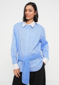 Ανοιχτό μπλε πουκάμισο με λευκό γιακά, με μακριά μανίκια και τσέπη στο στήθος, σε συνδυασμό με μπλε πλεκτό πουλόβερ δεμένο στη μέση.