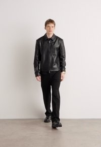 Giacca in pelle nera con zip frontale, abbinata a pantaloni e scarpe neri. Il modello è in piedi su uno sfondo neutro, guardando dritto.