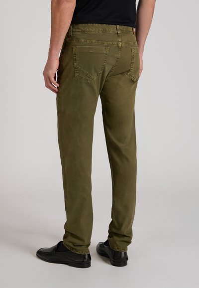 Man die olijfgroene broek en zwarte schoenen draagt, met zijn rug naar de camera toe staand tegen een effen achtergrond.