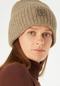 Brown ribbed knit beanie with a folded cuff and a leather patch featuring an embroidered design. Soft texture and neutral color.