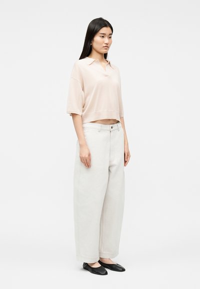 Γυναίκα που στέκεται φορώντας ανοιχτό ροζ φαρδιά κοντομάνικη μπλούζα, φαρδιά άσπρη παντελόνα και μαύρα μπαλαρίνες σε απλό ανοιχτό φόντο.