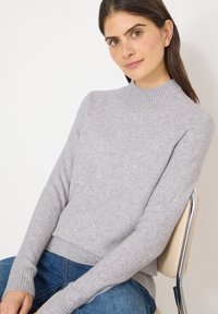 Hellgrauer Strickpullover mit einem hohen, gerippten Kragen und gerippten Bündchen. Weiche Textur, figurbetontes Design, kombiniert mit blauen Jeans.