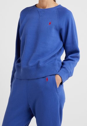 Person iført blå hættetrøje og matchende joggingbukser med røde broderede logoer, en hånd i lommen, ensfarvet baggrund.