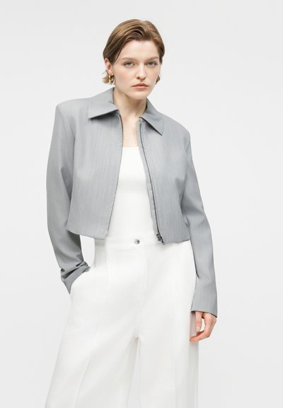 Γυναίκα με κοντά μαλλιά που φοράει χρυσούς κρίκους για σκουλαρίκια, γκρι cropped ζακέτα με φερμουάρ, λευκό τοπ και ψηλόμεσα λευκά παντελόνια, στέκεται με το ένα χέρι στην τσέπη.