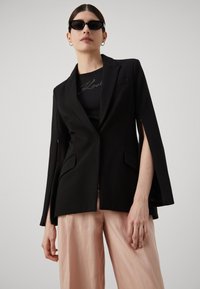 Čierny blazer s otvorenými rukávmi, strihom na mieru a bočnými vreckami. Nosený cez čierny top s strieborným logom a v páre s bledoružovými nohavicami.