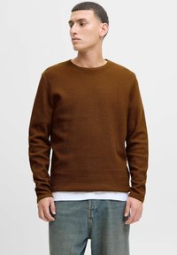 Brauner Waffelstrick-Pullover mit langen Ärmeln, Rundhalsausschnitt, lässige Passform, über einem weißen Hemd getragen, kombiniert mit hellblauen Jeans.