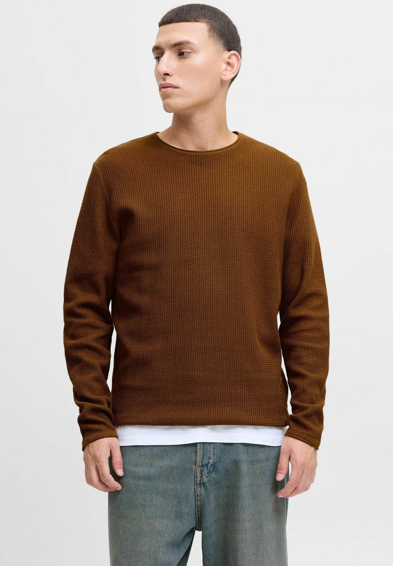 Brauner Waffelstrick-Pullover mit langen Ärmeln, Rundhalsausschnitt, lässige Passform, über einem weißen Hemd getragen, kombiniert mit hellblauen Jeans.