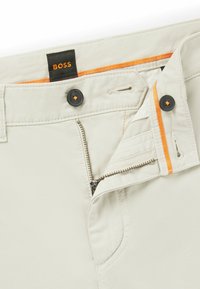 Beige bomuldsbukser med lynlås, med orange detaljer og mørke knapper. Mærket viser "BOSS" i taljen.