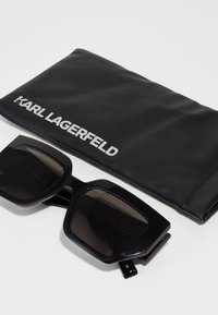 Lunettes de soleil noires avec verres sombres, dotées de montures épaisses ; accompagnées d'une pochette en cuir noir portant le logo "KARL LAGERFELD".