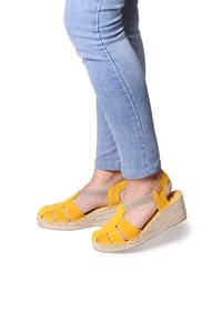 Personne portant un jean bleu clair et des sandales à talons compensés jaunes avec des semelles tressées, se tenant contre un fond blanc.