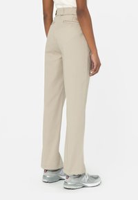 Pantalon beige à jambes larges et taille haute, avec des poches arrière et une texture lisse, associé à des chaussures de sport grises.
