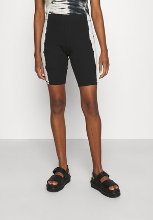 Persoon die zwart-witte tie-dye wielrennersshorts en zwarte sandalen draagt, staande tegen een witte achtergrond.