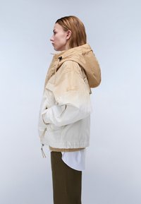 Kvinde i beige og hvid hættetrøje med hænderne i lommerne, hvid skjorte og olivengrønne bukser, vendt mod venstre mod en ensfarvet baggrund.