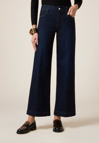 Los jeans de pierna ancha en azul oscuro cuentan con una cintura de media altura, bolsillos delanteros y traseros con costuras visibles.