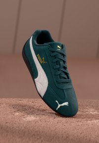 Teal sule sneakers valge ja kuldse aktsendiga, ümmargune varvas ja lame must kummitald. Logo on selgelt nähtav küljel.