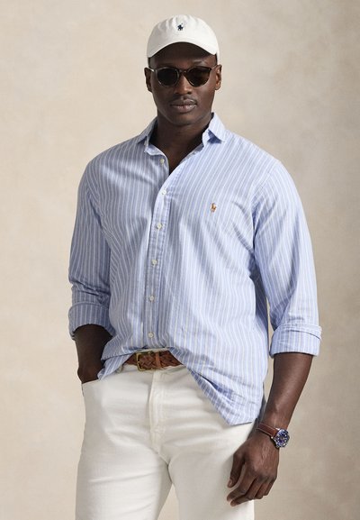 Camisa de botones de rayas azul claro con mangas largas, gorra blanca de béisbol, gafas de sol oscuras, cinturón tejido marrón y pantalones blancos.