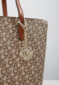 Sac fourre-tout en tissu beige avec un motif logo répétitif. Poignées en simili cuir marron avec ferrures métalliques dorées et un charm décoratif.