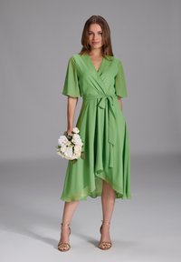 Robe verte à manches courtes et évasées, avec une taille à nouer et un ourlet plongeant. Associée à des sandales à brides dorées et un bouquet de fleurs blanches.