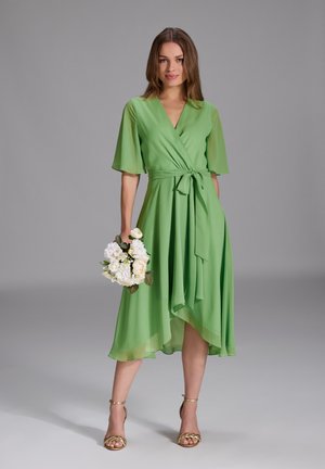 Grünes Wickelkleid mit kurzen, ausgestellt geschnittenen Ärmeln, einer taillierten Bindung und einem asymmetrischen Saum. Kombiniert mit goldenen Riemensandalen und einem Strauß weißer Blumen.