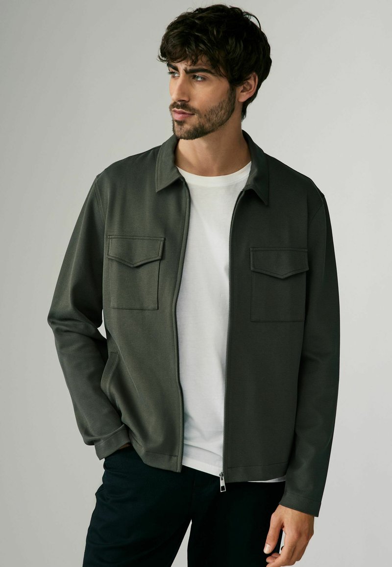 Chaqueta verde oscura con cremallera hecha de tela texturizada, con dos bolsillos en el pecho, usada sobre una camiseta blanca, combinada con pantalones oscuros.