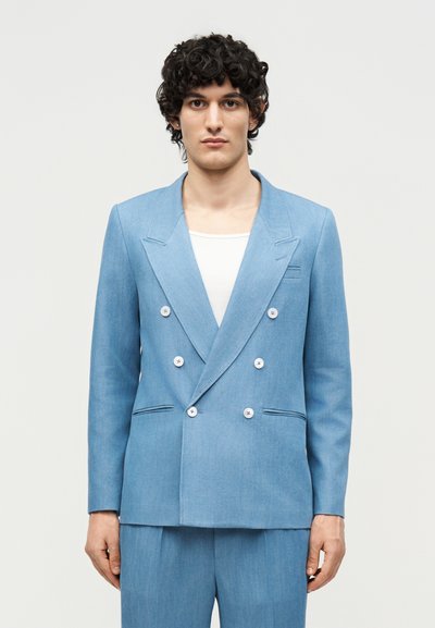Jeune personne aux cheveux noirs bouclés portant un blazer bleu clair croisé par-dessus un haut blanc sur un fond uni.