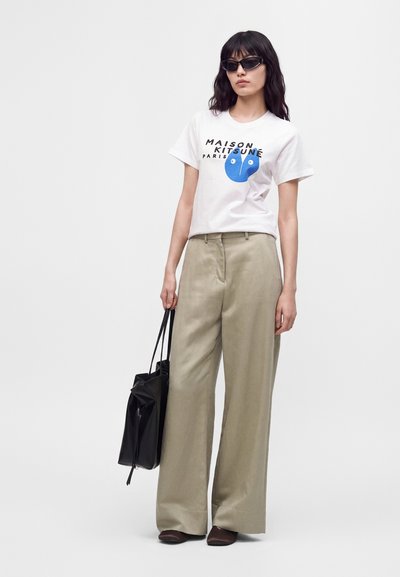 Γυναίκα με μαύρα γυαλιά ηλίου, λευκό μπλουζάκι με σχέδιο, μπεζ παντελόνι με φαρδιά μπατζάκια, κρατώντας μια μαύρη τσάντα ώμου σε απλό φωτεινό φόντο.