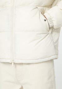 Chaqueta de plumas en suave tela crema, con cremallera completa, puños elásticos y bolsillos laterales con cierres a presión. Superficie texturizada y suave.