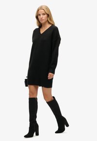 Vestido suelto negro de punto con escote en V, mangas largas y puños acanalados, combinado con botas altas de ante negras y un bolso pequeño.