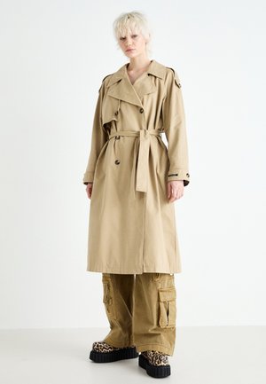 Beige Trenchcoat mit einem taillierten Gürtel, doppelt gereihten Knöpfen und Schulterklappen, kombiniert mit Cargo-Hosen und Plattformschuhen im Tierdruck.