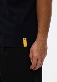 Μαύρο βαμβακερό t-shirt με κίτρινη ετικέτα στο στρίφωμα που φέρει κόκκινο κείμενο. Κοντά μανίκια, στρογγυλή λαιμόκοψη και άνετη εφαρμογή.