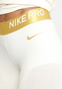 Las mallas Nike Pro cuentan con una cinturilla elástica dorada con el texto "NIKE PRO", un tejido blanco con detalles texturizados y un pequeño logotipo del swoosh de Nike en dorado.