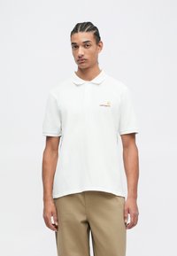 Jonge man met gevlochten haar, gekleed in een wit Carhartt poloshirt en beige broek, staande voor een effen witte achtergrond.