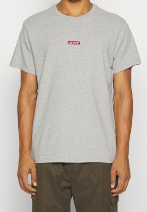 Mężczyzna ubrany w jasnoszary t-shirt z krótkim rękawem z małym czerwonym logo Levi's na piersi oraz ciemnooliwkowe spodnie typu cargo.