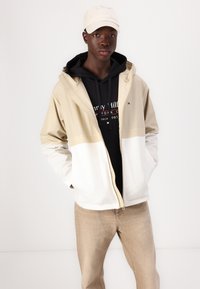 Beige and white color-blocked jacket with an adjustable hood, worn over a black hoodie. Beige cap, light brown textured pants.