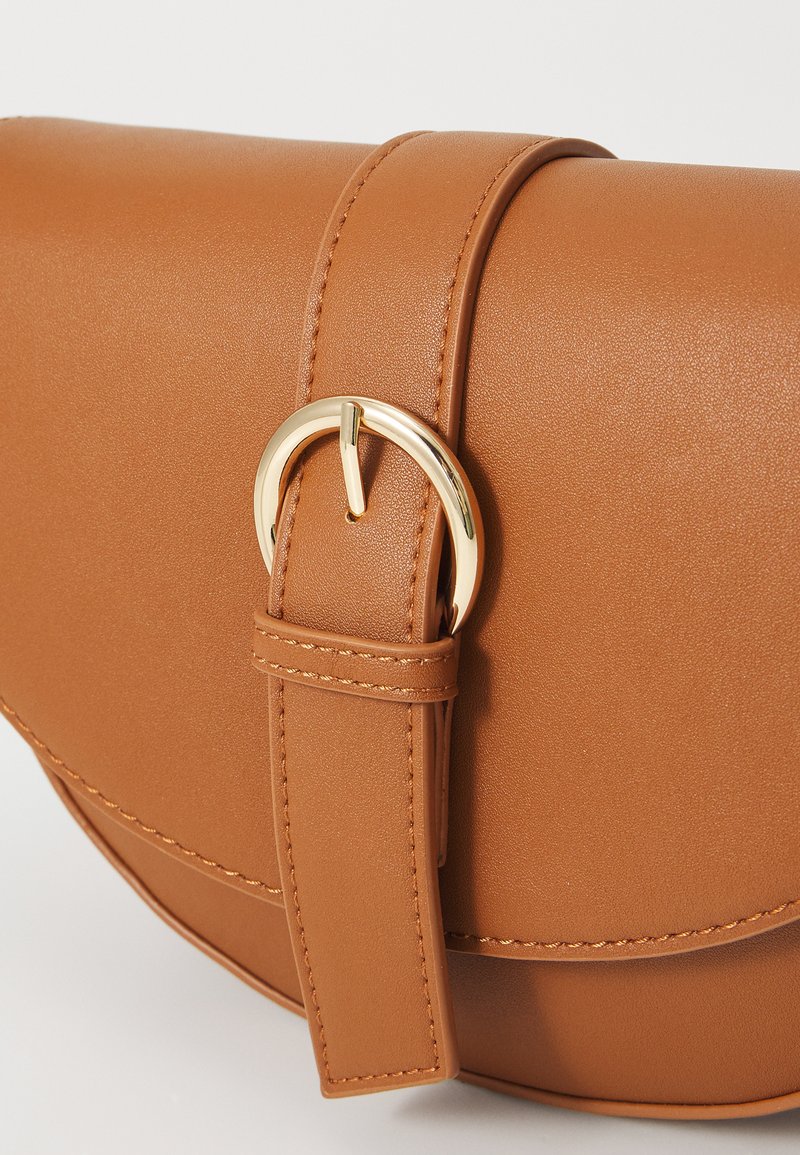 Sac en simili cuir marron de forme arrondie, avec une boucle en métal doré et des accents cousus sur la sangle et le corps.