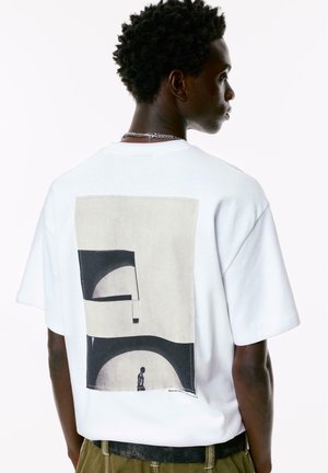 White cotton t-shirt featuring a large abstract graphic in black and beige on the back. Short sleeves, round neckline, relaxed fit.