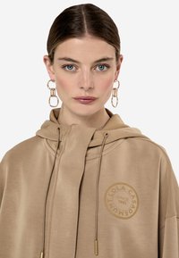 Sudadera beige hecha de una tela suave con un estampado de logo. Presenta una capucha con cordón y detalles en tono dorado.