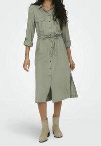 Abito camicia verde chiaro con maniche lunghe, chiusura con bottoni, due tasche sul petto e cintura in vita, caratterizzato da uno spacco laterale e tessuto testurizzato.