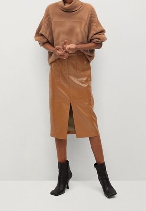 Pull en maille côtelée marron avec col montant associé à une jupe midi en cuir caramel dotée d'une fente avant et de deux poches avant. Bottines à talons noires.