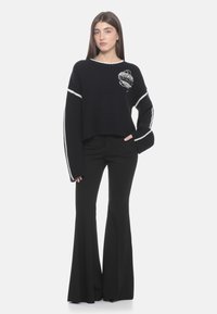 Navyblauer, kurzer Pullover mit geripptem Muster, ausgestattet mit weißen Streifen an den Ärmeln und einem grafischen Design. Kombiniert mit schwarzen Schlaghosen.