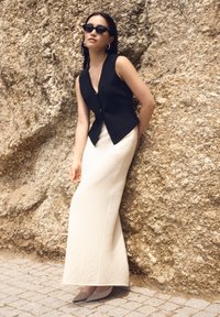 Crna bezrukavna majica s V izrezom, uparena s krem suknjom do tla i sivim cipelama s oštrim vrhom, protiv teksturirane stjenovite pozadine.